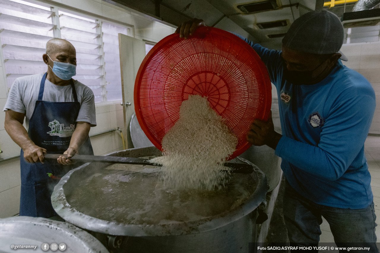 Dua sukarelawan menuang beras ke dalam kawah untuk dijadikan bubur. - Gambar oleh Sadiq Asyraf